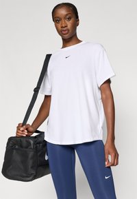 Λευκό κοντομάνικο T-shirt με μαύρο λογότυπο Nike, συνδυασμένο με μπλε κολάν. Το μοντέλο κρατάει μια μαύρη τσάντα duffel με σήμα Nike.