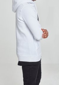 Witte capuchontrui met een kangoeroezak, geribde boorden en zoom. Draagt over een zwart T-shirt, gecombineerd met donkere broek. Soepele textuur.