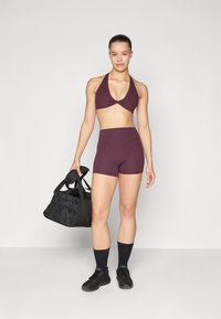 Bordo sportski-top i kratke hlače od rastezljivog materijala, s halter dizajnom i visokim strukom. Crna teretana torba s drškama u ruci.