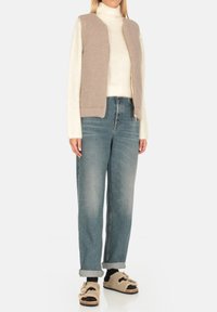 Gilet di maglia beige su un dolcevita bianco, abbinato a jeans denim blu con vestibilità rilassata e sandali in pelliccia nera e beige.