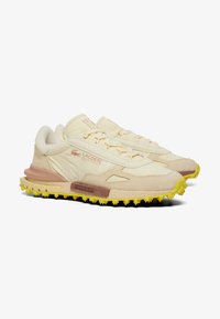 Zapatillas Lacoste con un upper de lona beige claro, detalles en ante, suela de goma amarilla y cierre de cordones con detalles en tono marrón contrastante.