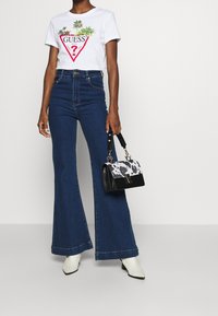 Bílá tričko s logem "GUESS" a palmovým potiskem, modré rozšířené džíny a černá kabelka s detailem kravského potisku. Bílé boty na podpatku.