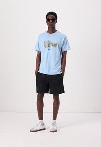 Lyseblå T-skjorte med grafisk trykk som sier "Santa Cruz", kombinert med svarte shorts og hvite sneakers. Modellen har på seg solbriller.