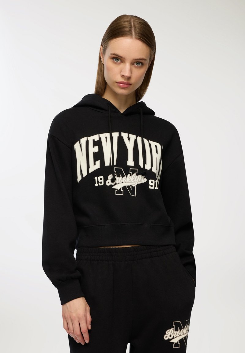Czarna krótka bluza z kapturem, wykonana z miękkiego materiału, z grafiką w kształcie łuku zatytułowaną „NEW YORK” oraz regulowanym kapturem. W połączeniu z czarnymi dresami.
