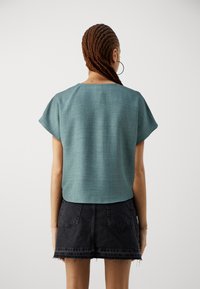 Frau mit geflochtenen Haaren, die ein türkisfarbenes Kurzarmoberteil und einen schwarzen Jeansrock trägt, von hinten vor einem schlichten Hintergrund betrachtet.