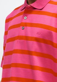 Ροζ και πορτοκαλί ριγέ πόλο μπλούζα με δύο κουμπιά και μικρή κέντηση "MN" στο στήθος, φορεμένη από ένα άτομο.