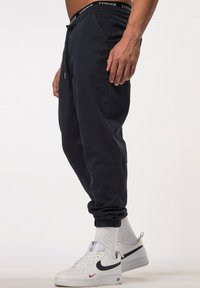 Marine joggingbroek met elastische boorden, een trekkoord in de taille en zijzakken. Draagt witte Nike sneakers en witte crew-sokken.