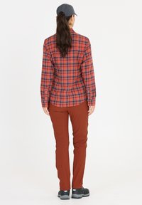 Punainen ja laivastonsininen ruudullinen nappipaita, pitkät hihat, yhdistettynä ruskeisiin slim-fit-housuihin ja mustiin kenkiin. Malli on kääntynyt poispäin, päässään hattu.