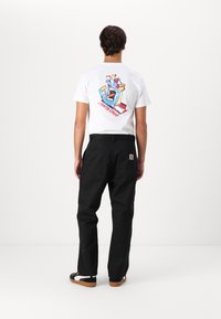 Weißes T-Shirt mit einem bunten Grafikdesign einer Hand auf dem Rücken; schwarze Hose mit zwei Gesäßtaschen, getragen mit schwarzen und beigen Schuhen.