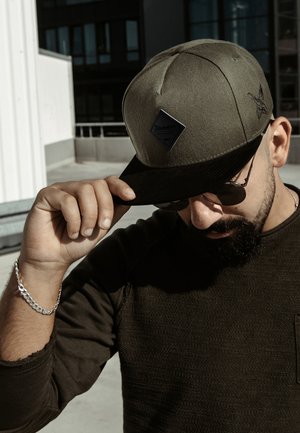 Gorra verde oliva con visera negra, que presenta un parche en forma de diamante y detalles bordados; llevada con gafas de sol y una camiseta oscura texturizada.