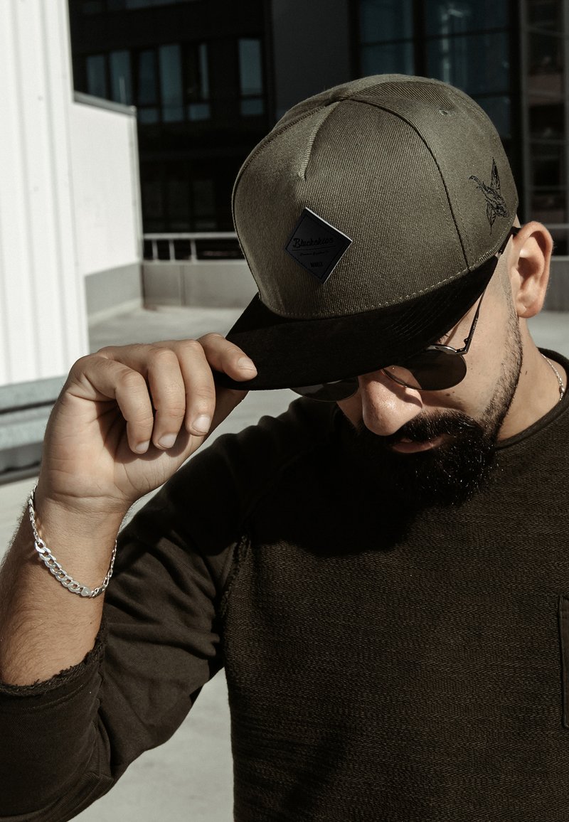 Cappello verde oliva con visiera nera, dotato di una toppa a forma di diamante e dettagli ricamati; indossato con occhiali da sole e una camicia scura con texture.