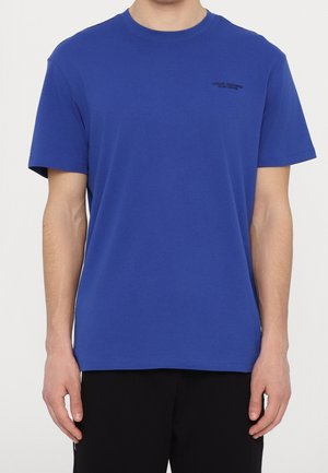 Homme porte un t-shirt bleu uni à manches courtes avec un petit texte noir sur la poitrine gauche et un pantalon noir, debout devant un fond blanc.