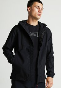 Fekete kapucnis dzseki cipzárral, elülső zsebekkel és elasztikus mandzsettákkal. Diszkrét logó részlet jellemzi, sima, vízálló textúrával.