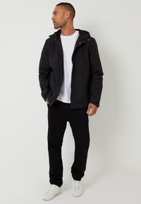 Sort zip-jakke med hætte, der har en glat overflade og sidelommer, båret over en hvid t-shirt og sorte bukser med hvide sneakers.