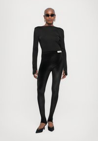 Black long-sleeve top and high-waisted leggings made of a soft, shiny material. Model wears black pointed-toe heels and sunglasses.
