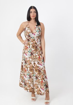 Kobieta o długich ciemnych włosach, ubrana w bezrękawową, kwiecistą suknię maxi z głębokim dekoltem i białe sandały na obcasie, stojąca z jedną ręką na biodrze.
