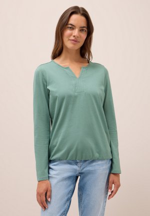 Haut à manches longues en coton vert doux, avec un col en V. Présente une patte de boutonnage au niveau du col et une coupe décontractée. Associé à un jean bleu clair.