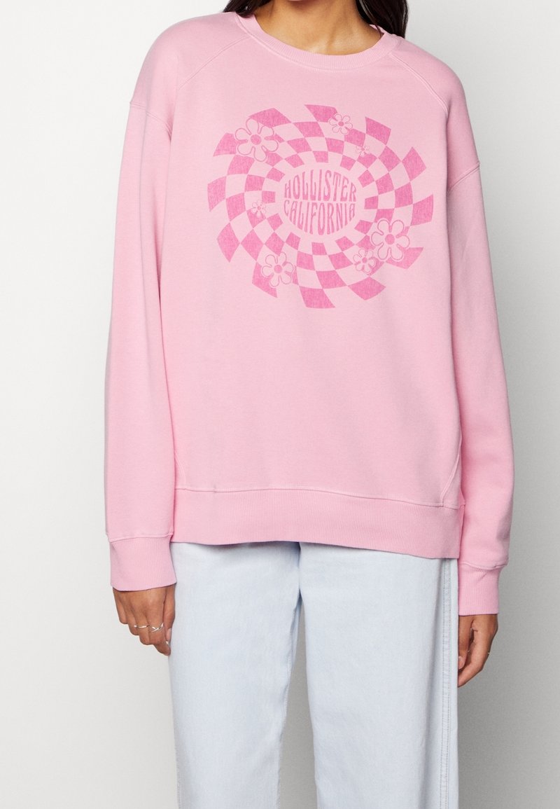 Rosa sweatshirt med rutmönster och blommiga detaljer samt texten "Hollister California" i mitten. Mjuk tyg, crewneck-design.