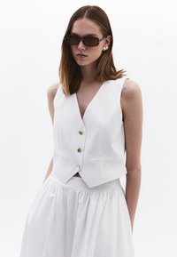 Weißes ärmelloses Oberteil mit tiefem V-Ausschnitt, goldenen Knöpfen und seitlichen Taschen, kombiniert mit einem weißen, fließenden Rock. Glatte Textur und taillierte Passform.