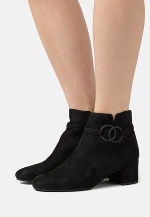 Schwarze Wildleder-Ankle Boots mit Blockabsatz, ausgestattet mit einem Riemen und einem kreisförmigen Akzentdetail an der Seite. Glatte Textur und minimalistisches Design.