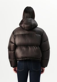 Giacca a vento marrone con cappuccio, caratterizzata da una texture lucida e trapuntata con polsini elasticizzati. Indossata con jeans in denim scuro.