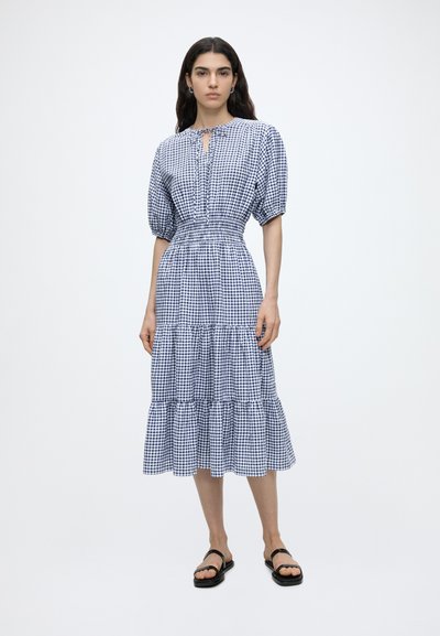Femme debout portant une robe midi vichy bleue et blanche à manches bouffantes et des sandales noires à bouts ouverts sur un fond blanc uni.