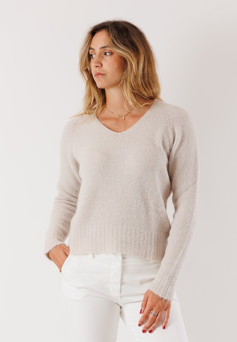 Maglione panna a V in morbido tessuto testurizzato con polsini e orlo a coste, indossato sopra pantaloni bianchi.