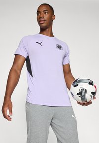 Camiseta deportiva de color púrpura claro con acentos y logo en negro, combinada con pantalones grises. El modelo sostiene un balón de fútbol con un diseño estampado.