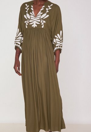 Femme portant une longue robe vert olive avec des broderies de feuilles blanches sur la poitrine et les manches, debout contre un fond clair uni.