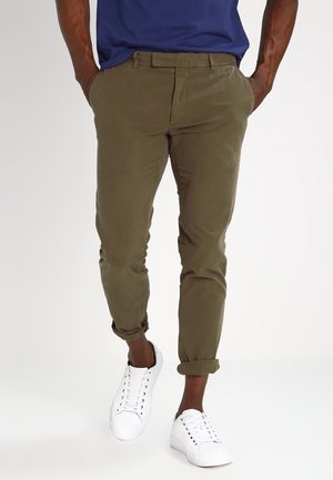 Man draagt olijfgroene opgerolde chino's gecombineerd met witte sneakers met veters en een marineblauw overhemd, handen in de zakken, staand op een witte achtergrond.