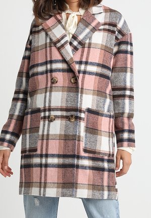 Žena v dvouřadém kabátu délky ke kolenům s velkým kostkovaným vzorem v růžových, hnědých, bílých a námořnicky modrých tónech, přes světle modré džíny.