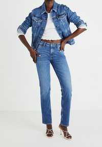 Denim-takkie i medium blå med to brystlommer og let søm. Hvid crop top og lige ben blå jeans parret med hvide hæle.