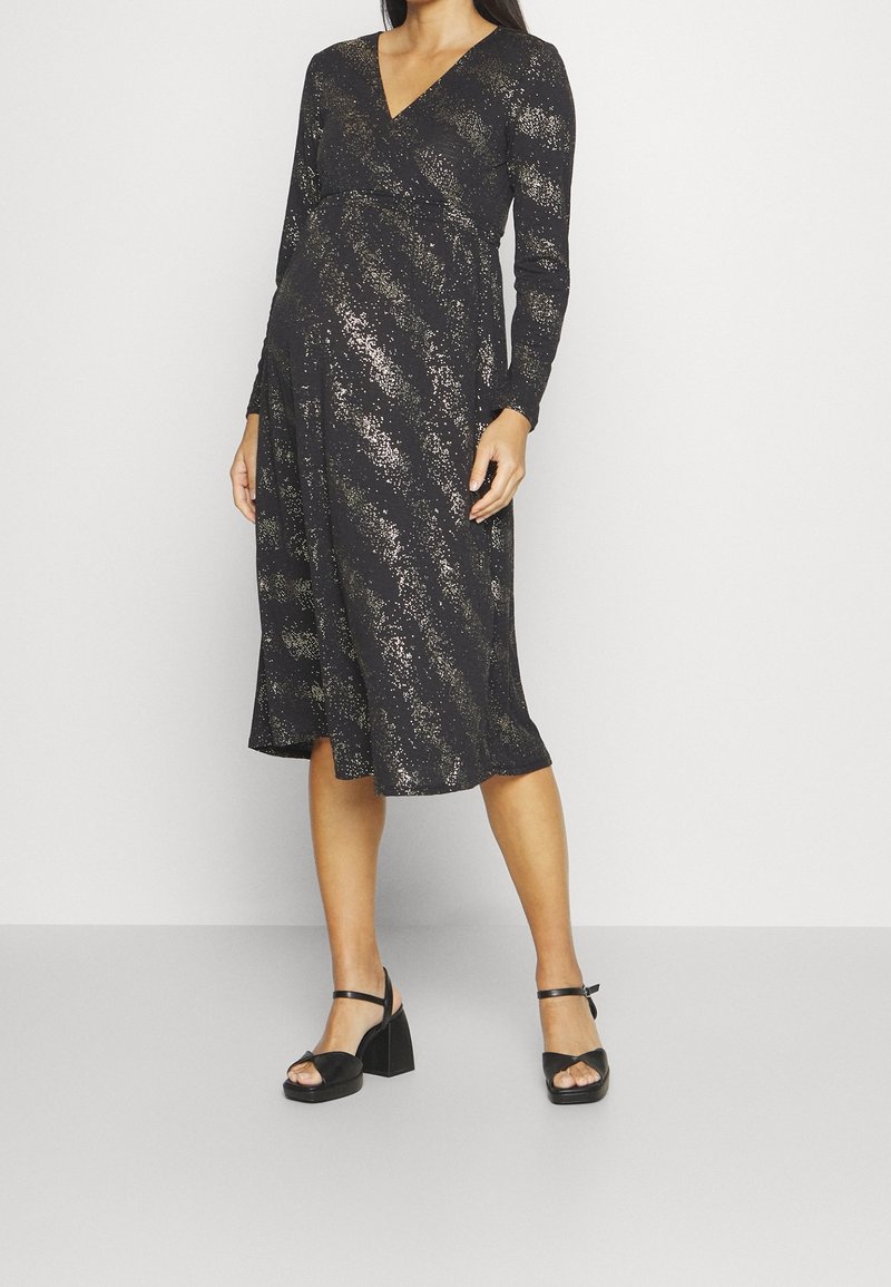 Svart långärmad gravidklänning med V-ringning och guldprickat mönster, med en omslag design och flödande kjol. Stiliserad med svarta blockklacksandaler.