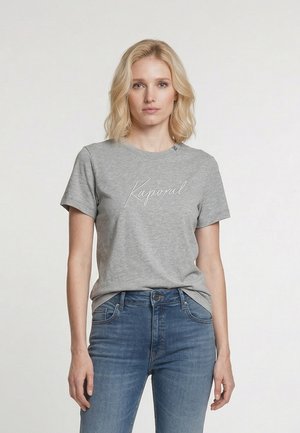 Blond vrouw die een grijs Kaporal t-shirt en blauwe spijkerbroek draagt, staand tegen een effen witte achtergrond met een neutrale gezichtsuitdrukking.