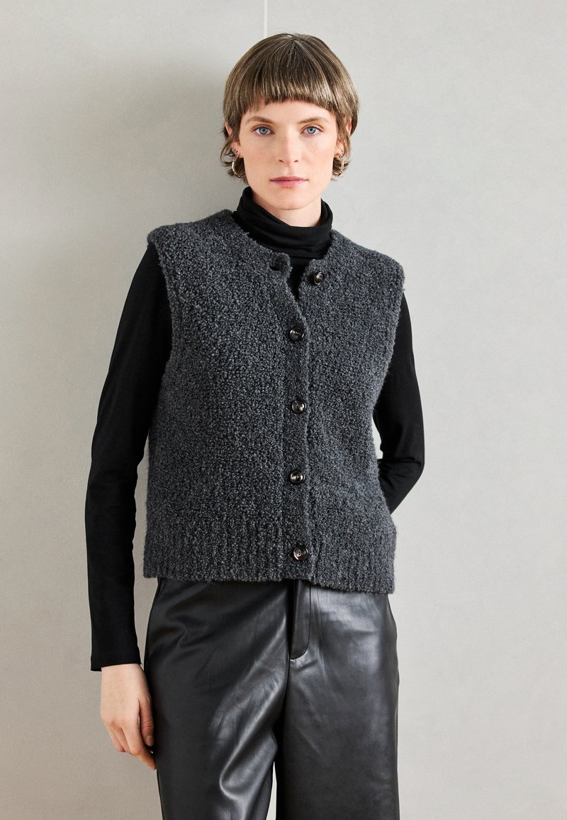 Hallitusseente struktuuriga hall kootud vest, millel on ümmargune kaelus ja esipainikud, kantakse musta kõrge kaelusega pluusi peal. Nahkpüksid täiustavad väljanägemist.