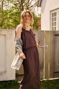 Femme souriante en robe marron sans manches et lunettes de soleil tenant une veste grise, debout à l'extérieur près d'une porte en bois par une journée ensoleillée.