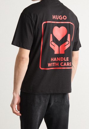 Person iført sort T-shirt med rød grafik af hænder, der holder et hjerte, og teksten "HUGO HANDLE WITH CARE" på bagsiden.