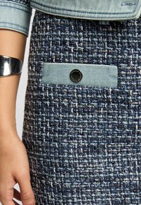Jeansrok met een gestructueerde marineblauwe weving, voorzien van een lichtblauwe denim zak en een zwarte knoopdetail. Glad, gestructureerd stof.