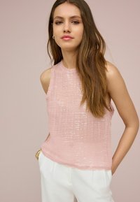 Top roz fără mâneci, dintr-un material texturat cu paiete, având un guler înalt și o croială lejeră, asortat cu pantaloni albi.