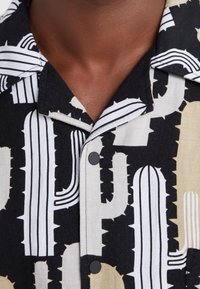 Chemise à manches courtes noire, blanche et beige, ornée d'un motif de cactus et de boutons noirs. Fabriquée en tissu léger avec une finition texturée.