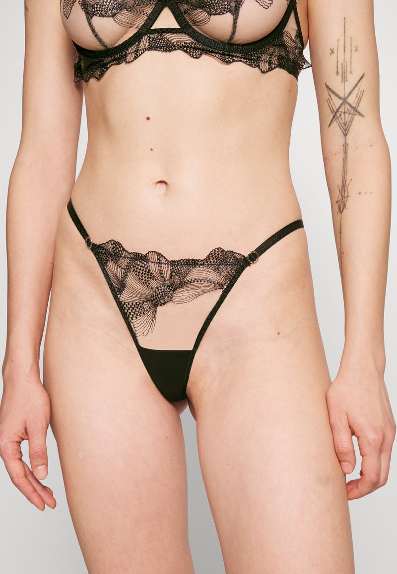 Conjunto de lingerie em renda preta com padrões florais intrincados, parte superior em malha transparente e fio dental de cintura baixa com detalhes em metal dourado.