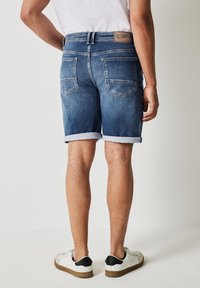 Jasnobłękitne szorty dżinsowe z podwiniętym dołem, tylnymi kieszeniami oraz brązową skórzaną naszywką z logo przy pasku. Gładka tekstura.