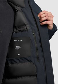 Marine foretaksjakke indvendigt med quiltet polstring, en sort lap med hvid tekst, og en lynlåslomme for ekstra opbevaring.