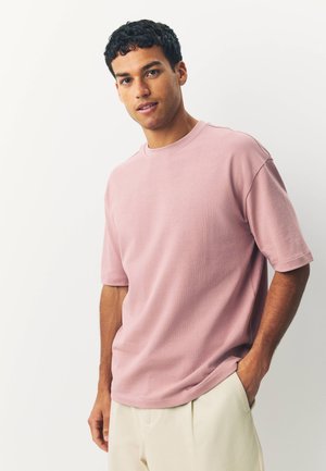 Fiatal férfi rövid göndör hajjal, világos rózsaszín túlméretezett pólót és bézs nadrágot visel, egyik kezét a zsebébe dugva áll.