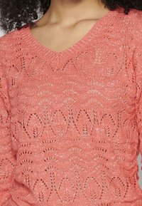 Pull en maille corail avec col en V, motif ajouré complexe et léger scintillement, présentant un design texturé et léger.