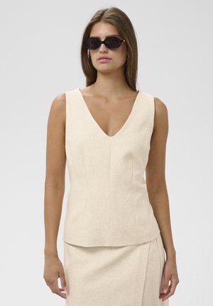 Femme portant un haut beige sans manches en V, une jupe assortie et des lunettes de soleil noires œil-de-chat sur un fond gris uni.