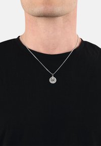 Silversmyckeshalsband med en rund berlock med intrikata detaljer, hängande på en tunn kedja, bärs över en svart rundhalsad tröja.