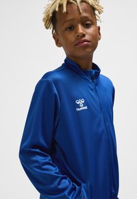 Blauer Reißverschluss-Pullover aus glattem Stoff mit hohem Kragen und dem "hummel"-Logo auf der linken Brust. Lange Ärmel mit eng anliegenden Bündchen.