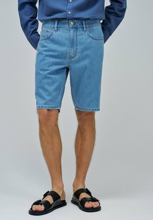 Un homme portant un short en jean bleu clair associé à des sandales noires à boucle et une chemise bleue à manches longues rentrée.
