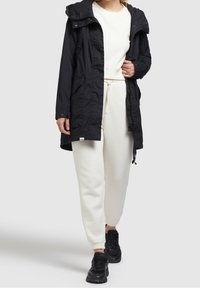 Svart parkas med huva, med knapp- och dragkedjestängning framtill, sidofickor och justerbara snoddar i midjan. Matchas med vita joggers.